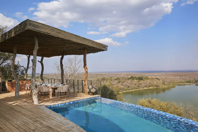 Luxury infinity pool deck with thatched pergola, set table, overlooking savanna lake under blue skies