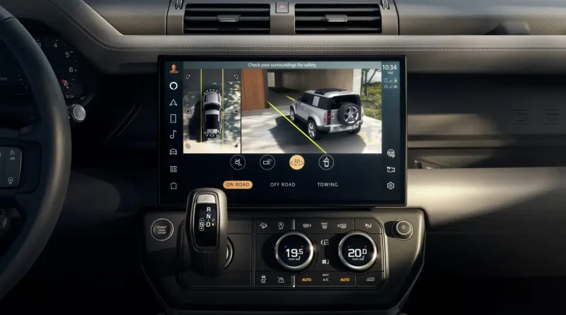 Land Rover Defender dashboard with central screen displaying rear camera view of white Defender SUV in garage.