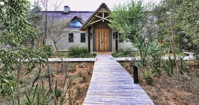 Monwana Game Lodge entrance: thatched-roof building with wooden door, flanked by trees, leading via boardwalk path in bushveld.