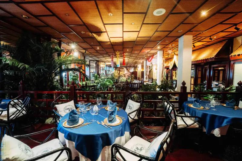 Elegant Hilton Malta restaurant interior with wooden ceiling, lush plants, blue-clothed tables set with glassware.