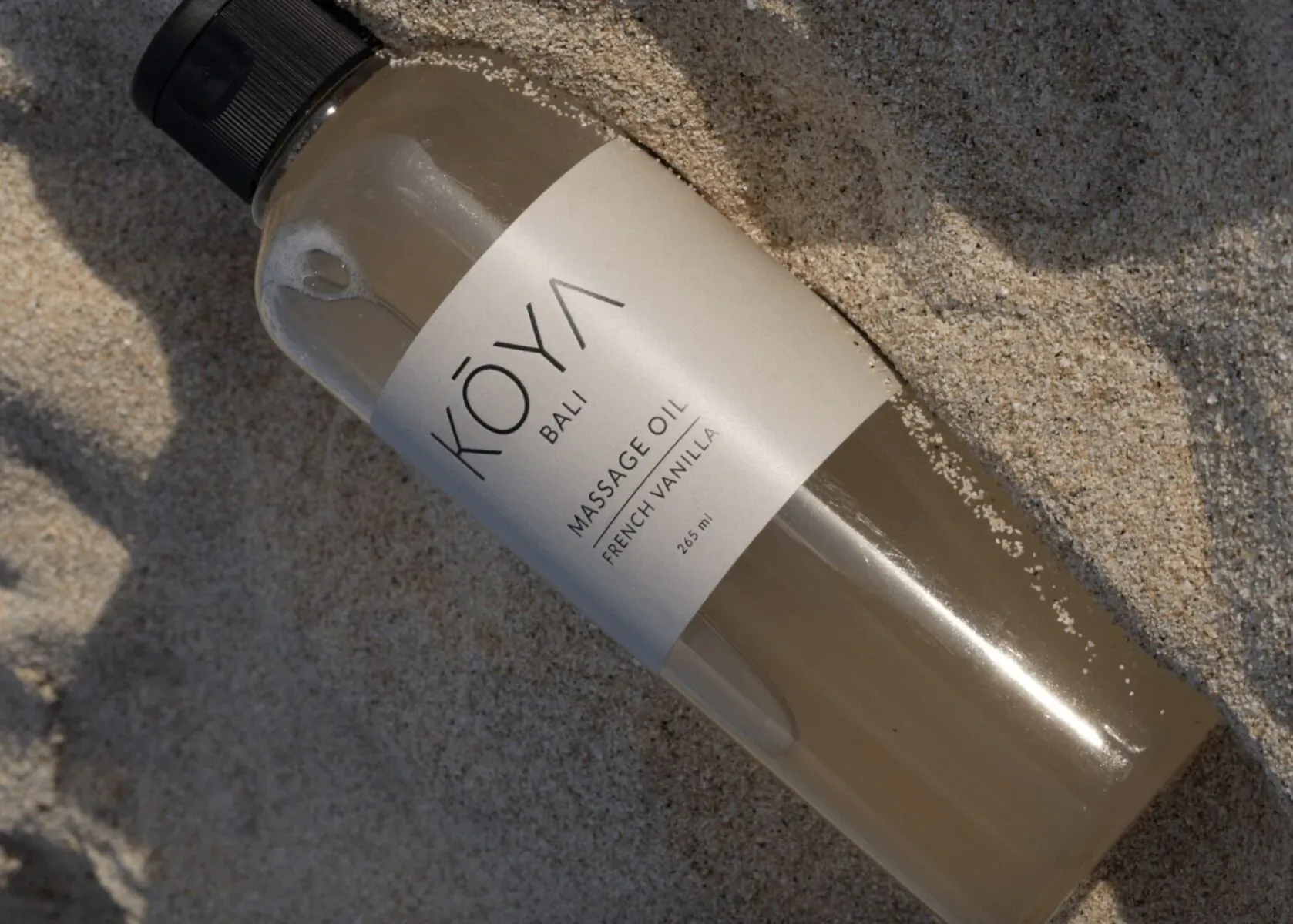 Koya Massage Vanilla Oil bottle lying on Bali beach sand, translucent with white label.