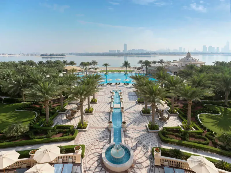 Aerial view of Raffles The Palm Dubai's luxurious infinity pool, palm trees, fountain, and Dubai skyline across the water.