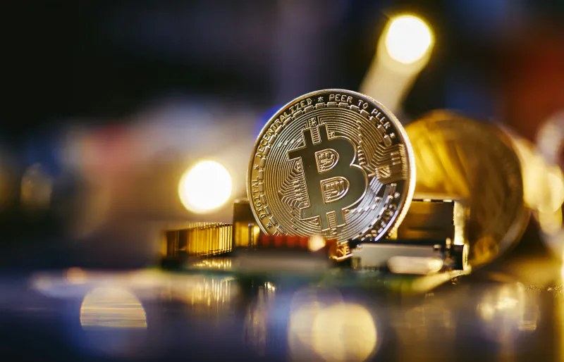 Shiny gold Bitcoin coin with Bitcoin symbol on a glowing circuit board, warm bokeh lights in background.