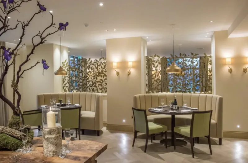 Elegant dining area at Careys Manor Hotel with curved cream booths, round table set with wine glasses and candle, gold leaf screens, and potted branches.