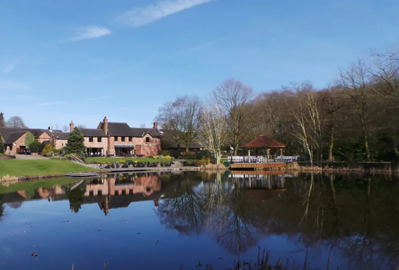 Moddershall Oaks Spa: brick manor house, gazebo by reflective pond amid bare trees under blue sky.