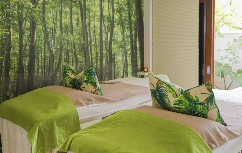 Two spa beds with green palm pillows and blankets in room with lush forest mural wallpaper