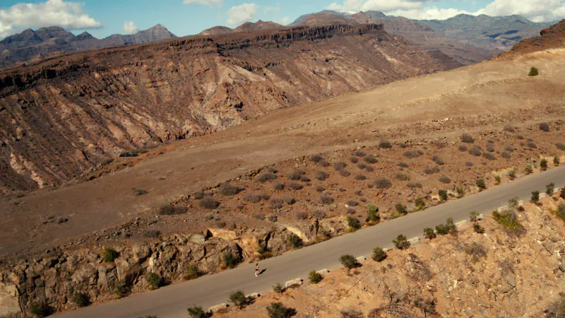 Aerial view of a winding road flanked by sparse bushes in a vast, rugged desert canyon with layered mountains under a partly cloudy sky.