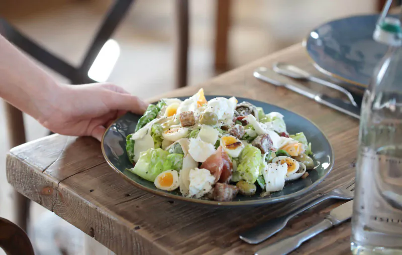 Hand serving Cobb salad with lettuce, eggs, ham, feta on blue plate at rustic wooden table with water bottle
