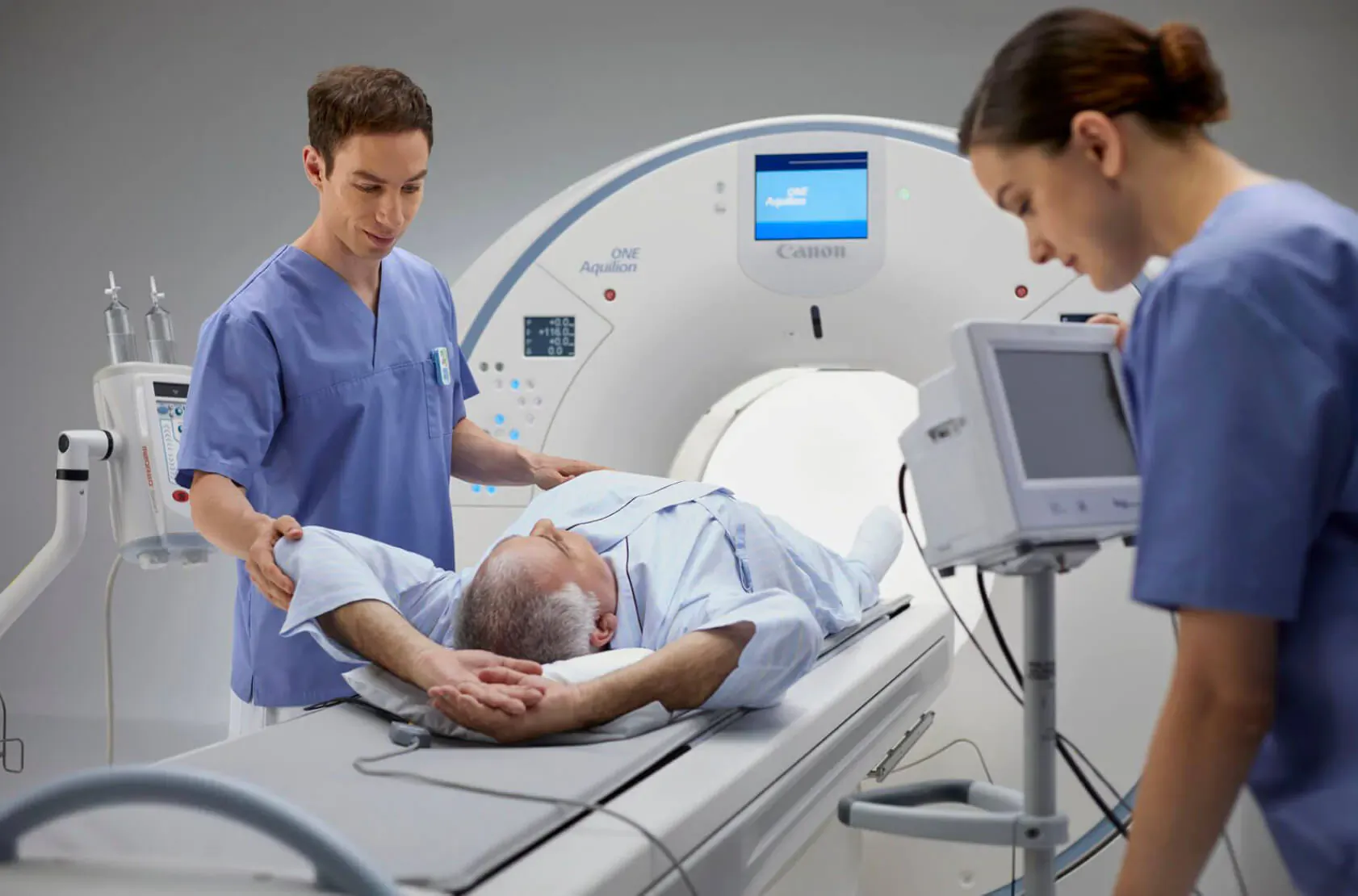 Two nurses in scrubs assist elderly male patient lying on MRI scanner bed, monitors visible.