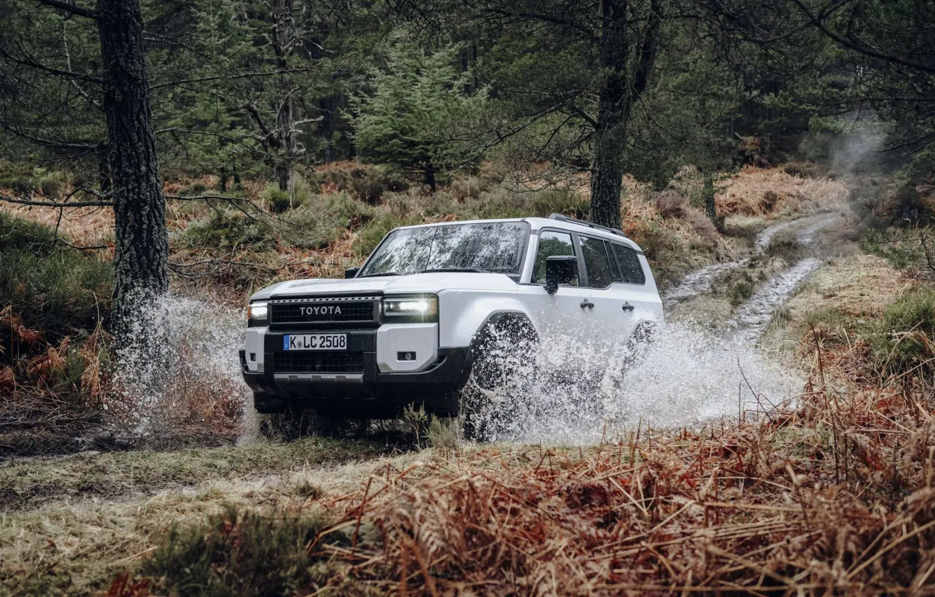 White Toyota Land Cruiser with 'TOYOTA' grille drives through splashing water on forested off-road trail