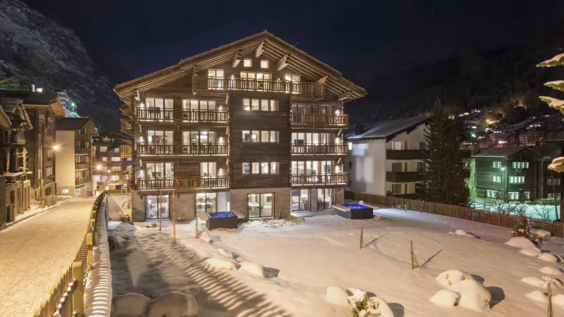 Luxury wooden ski chalet at night in snowy European alpine resort, surrounded by mountains and lights.