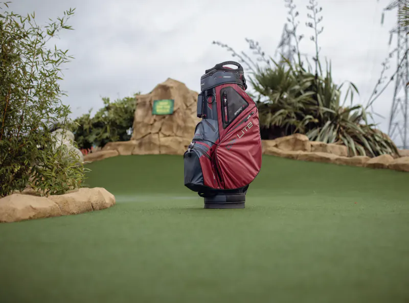Black and red John Deere golf bag standing on lush green mini-golf course amid rocks and plants
