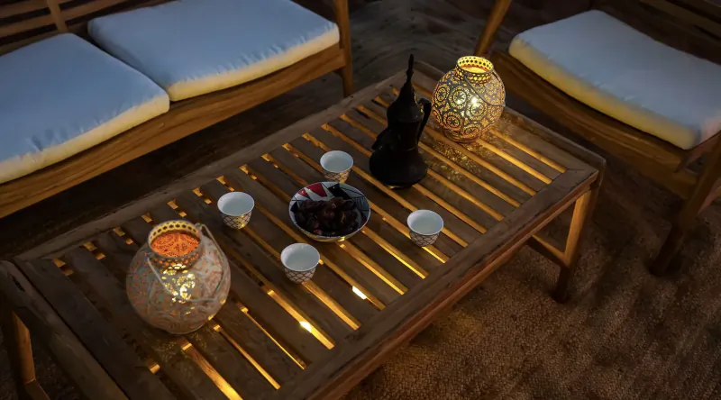 Wooden outdoor table with lit ornate lanterns, black statue, dates in bowl, and white teacups at dusk.