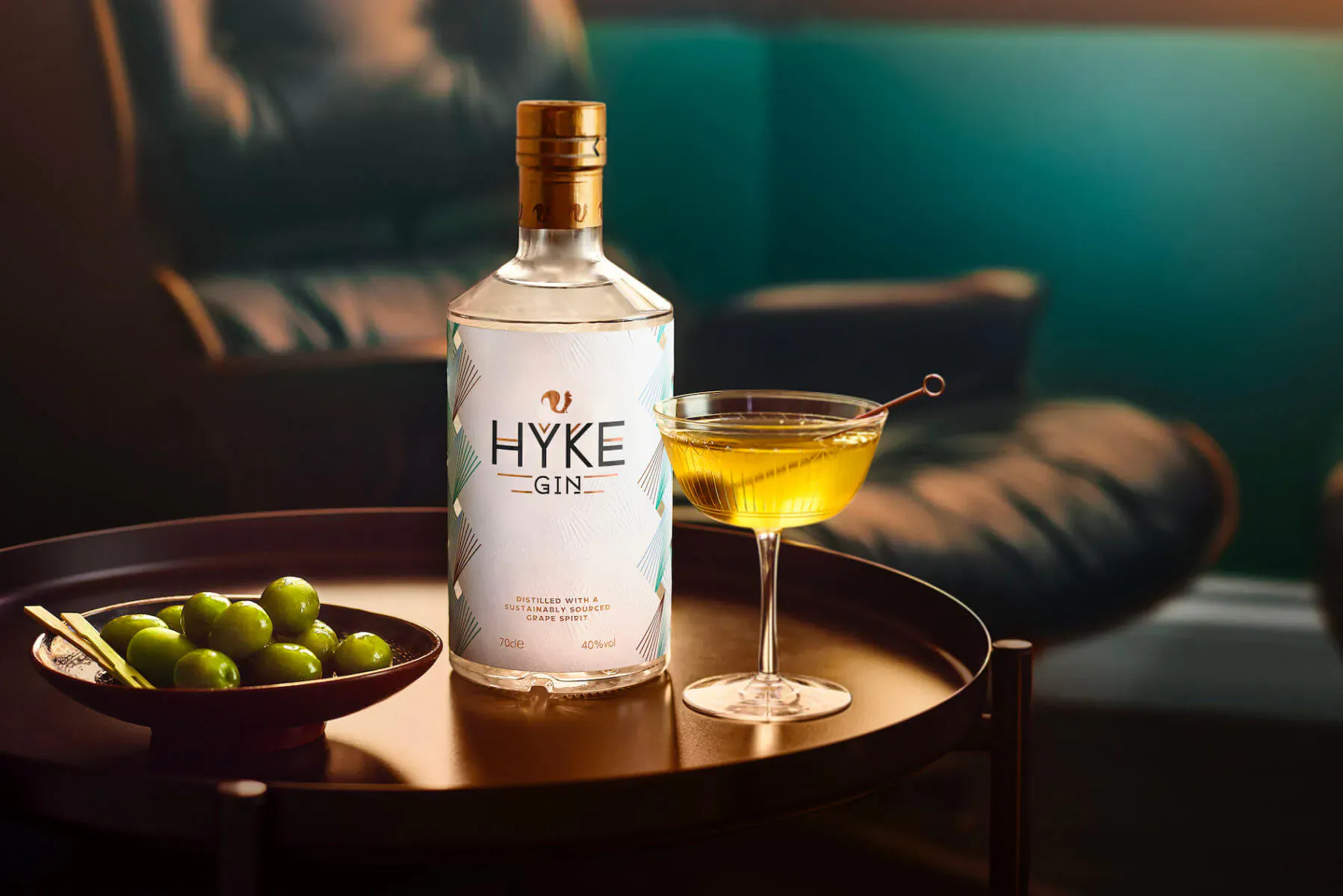 HYKE Gin bottle with yellow cocktail in martini glass and green olives on tray beside leather chair, teal background.