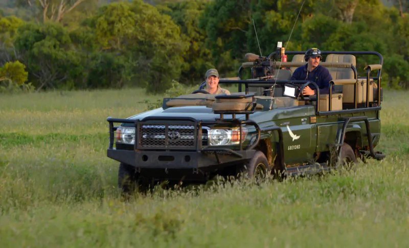 Two men in caps drive a green Land Cruiser safari vehicle through grassy savanna in Kruger National Park.