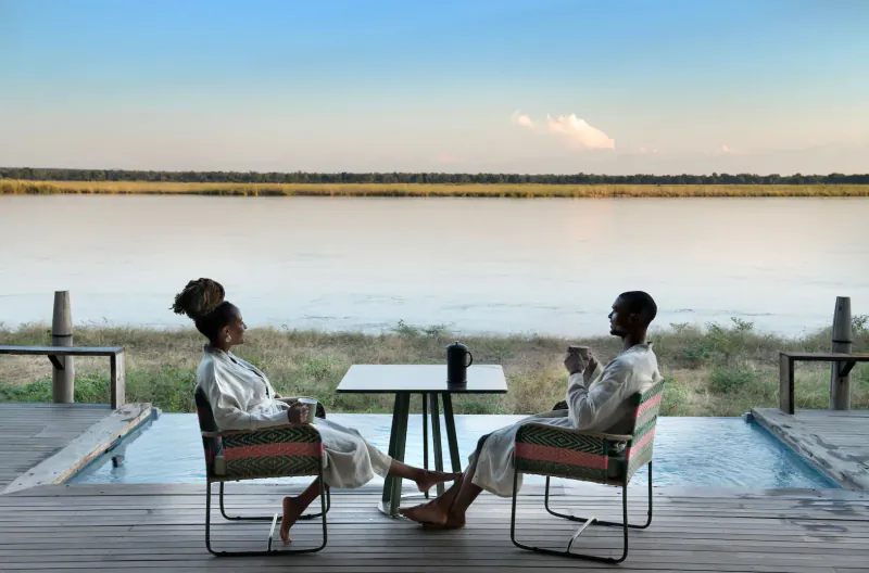 Black couple in white robes, sipping drinks at low table on deck overlooking serene river at Lolebezi luxury safari lodge.