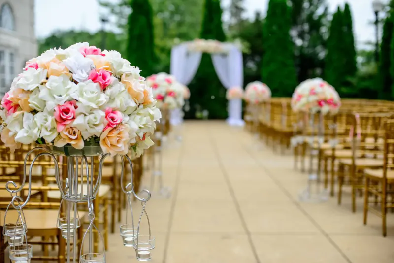 Outdoor wedding aisle with white floral arch, pink and white flower stands, gold chairs, and greenery backdrop