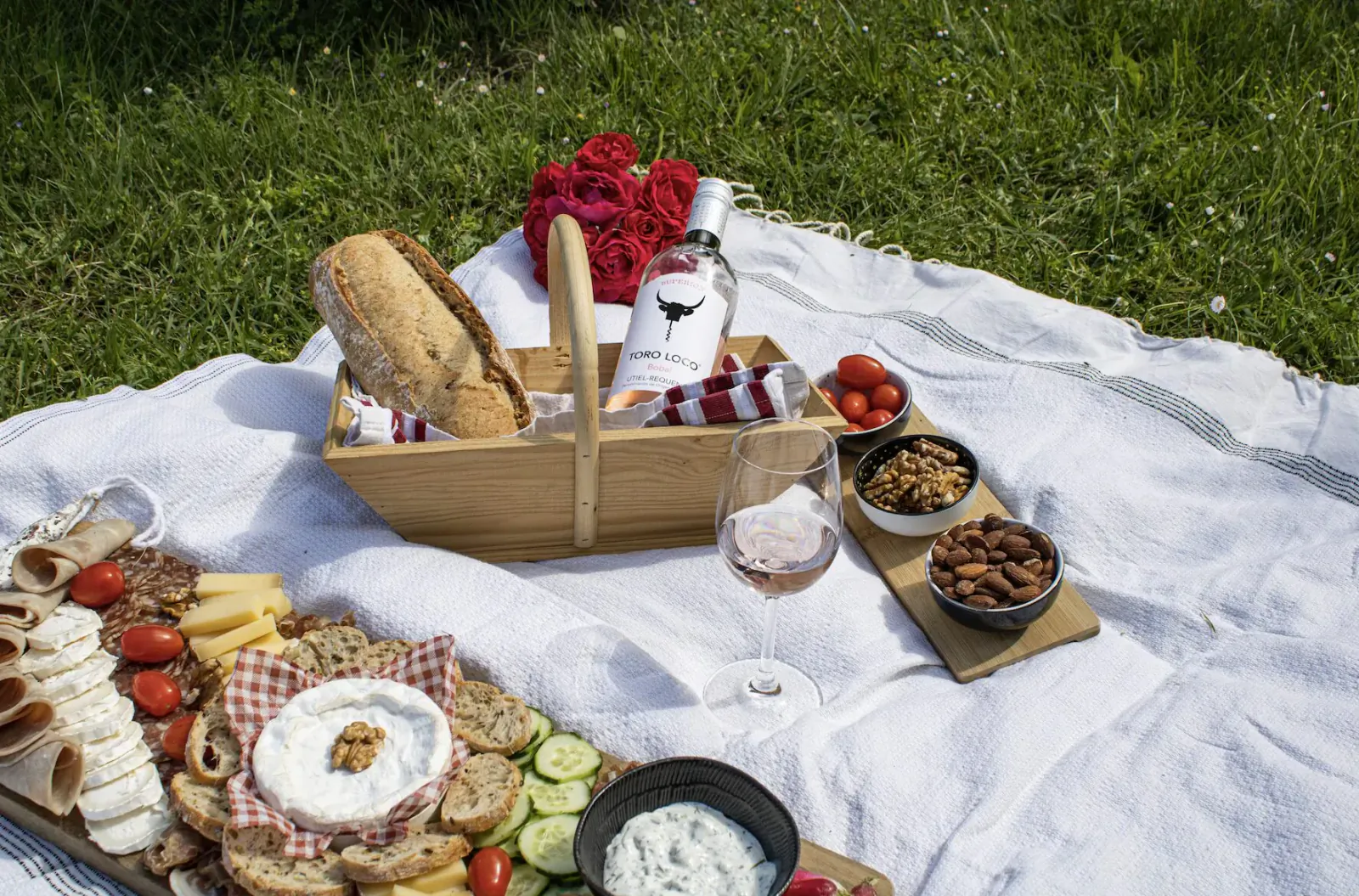 Picnic basket with baguette, charcuterie board, rosé wine glasses and bottle, nuts, tomatoes, red roses on checkered blanket in grass