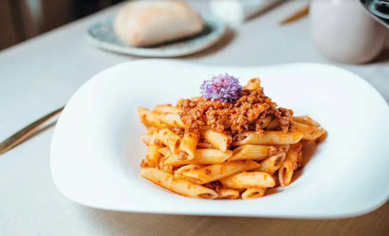 Plate of penne pasta topped with meat sauce and purple garnish, on table with bread roll at Italian restaurant.