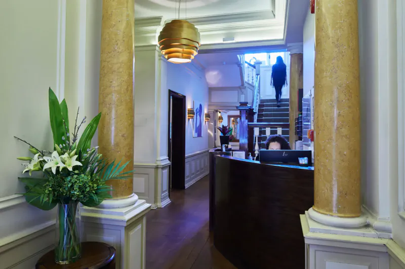 Elegant hotel lobby with curved reception desk, potted lilies, golden columns, pendant lamp, and person on stairs