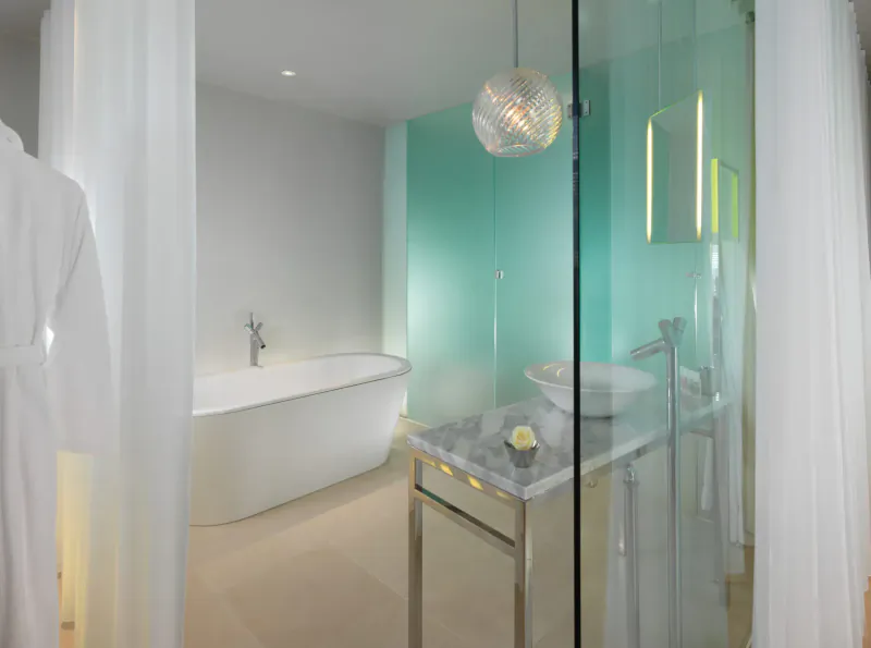 Modern luxury hotel bathroom with freestanding white bathtub, glass sink vanity, teal wall, pendant light, and sheer white curtains