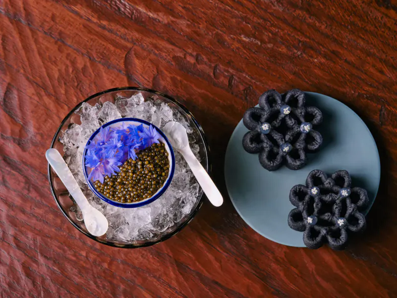 Overhead view at AngloThai: blue edible flowers on gray plate beside glass bowl of blue caviar on ice with spoons on wood table