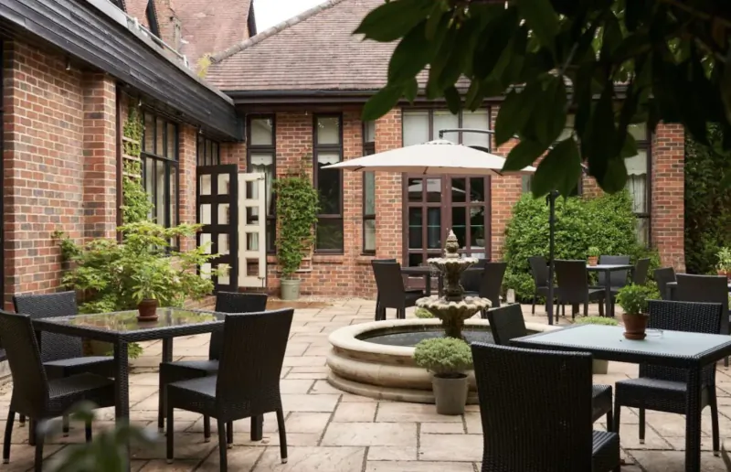Elegant outdoor courtyard at Careys Manor Hotel with black chairs, tables, fountain, plants, and umbrellas amid brick buildings.