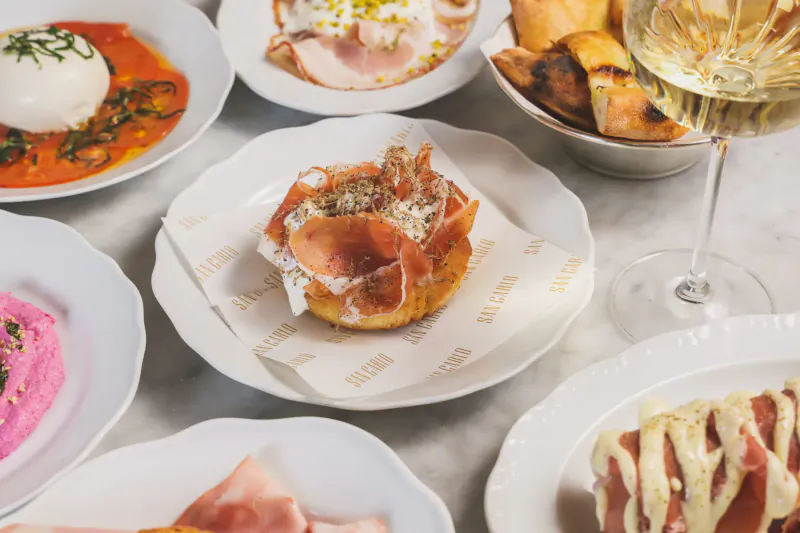 Assortment of Sicilian dishes on white plates: burrata, prosciutto, hummus, topped with wine glass