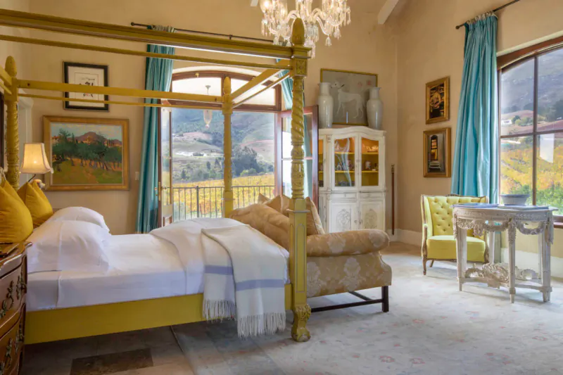 Luxurious Franschhoek bedroom with yellow four-poster bed, balcony view of vineyards, chandelier, and art.