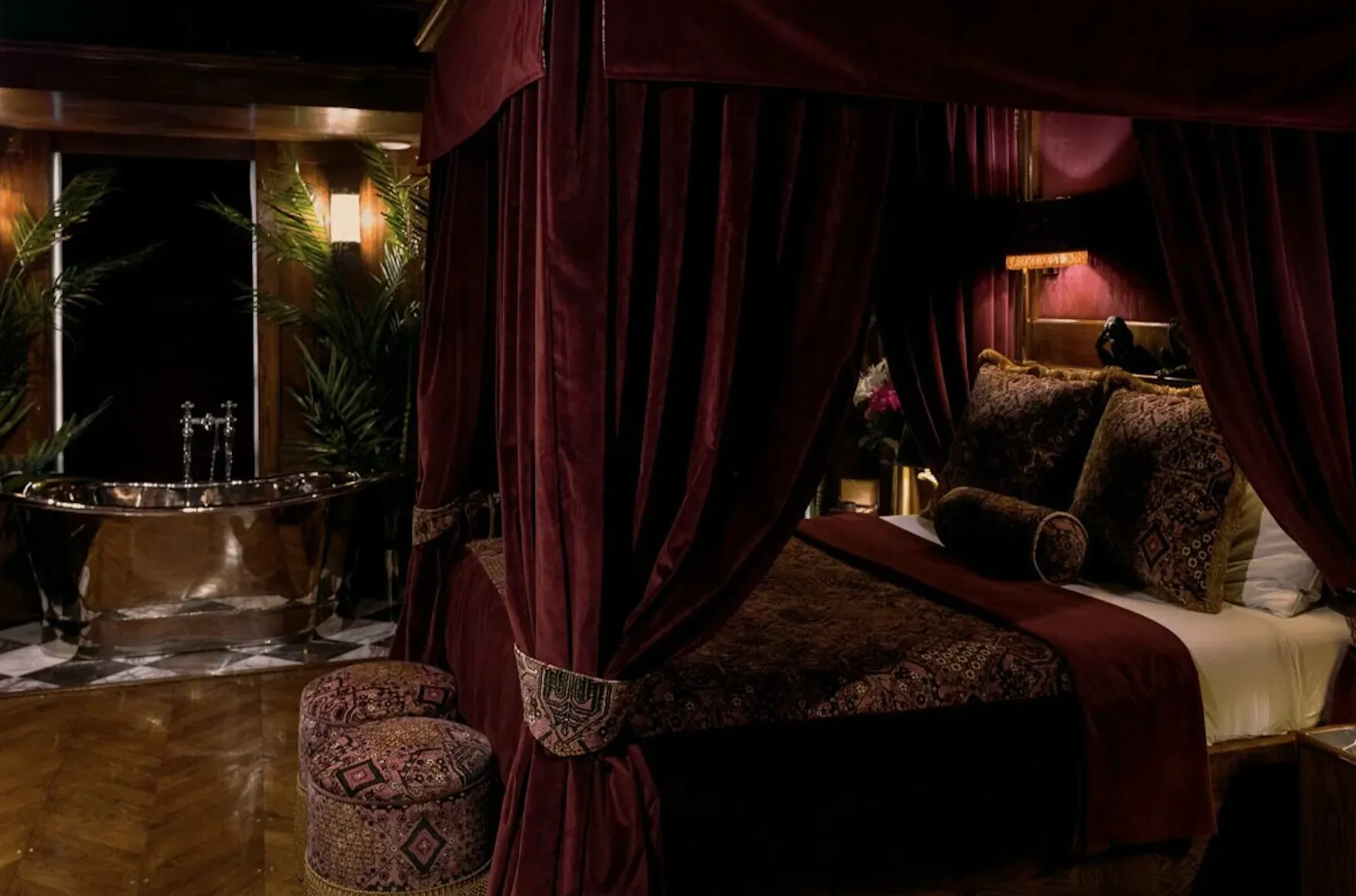 Luxurious burgundy four-poster bed and clawfoot bathtub in opulent Edinburgh hotel room with plants and warm lighting.