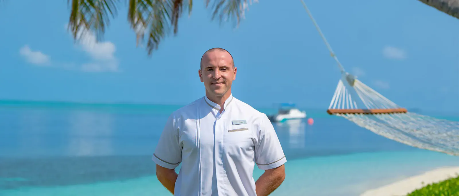 Amir Badr, smiling in white uniform, stands arms crossed at Hideaway Beach Resort & Spa with hammock, palms, and turquoise sea.