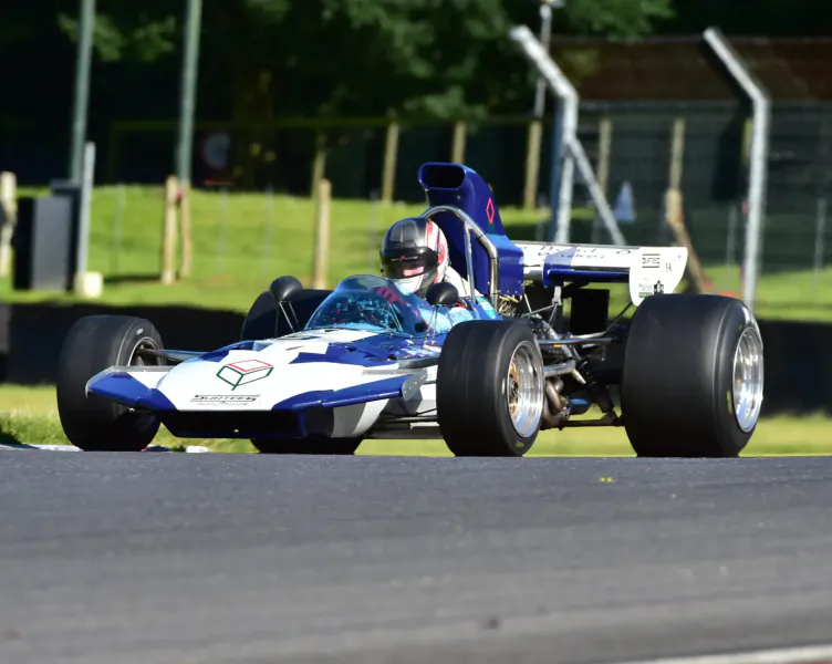 Blue and white Lotus racing car with helmeted driver speeding on track at London Concours 2024 event, surrounded by barriers and trees.