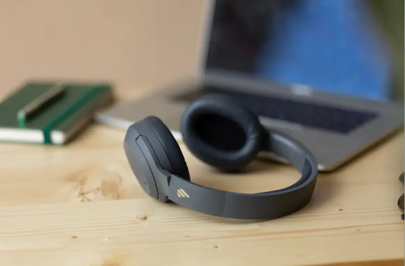 Edifier W820NB gray over-ear headphones beside open laptop and green notebook on wooden table