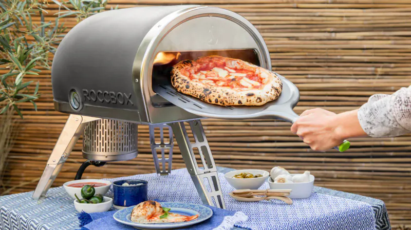 Gozney portable pizza oven with open door, woman using peel to remove hot margherita pizza, outdoor patio setting with toppings table.