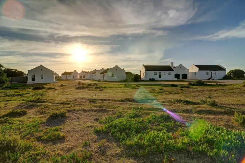 Row of white cottages in grassy field at De Hoop Preserve, sunset with sun rays and cloudy sky