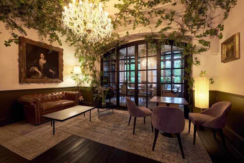 Elegant Can Bordoy hotel lounge with ivy-draped chandelier, portrait painting, leather sofa, velvet chairs, and arched garden doors.