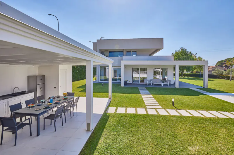 Modern white luxury villa in Sicily with covered outdoor dining table set on lawn under pergola, sunny day.