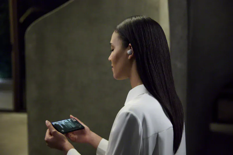 Asian woman in white shirt holds phone, wearing Sony WF-1000XM6 wireless earbuds, side profile in dim setting