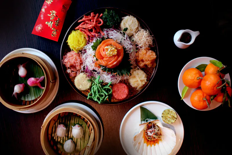 Top-down view of vibrant Chinese dim sum platter with dumplings, salmon flower, veggies, oranges, and red envelope on black table.