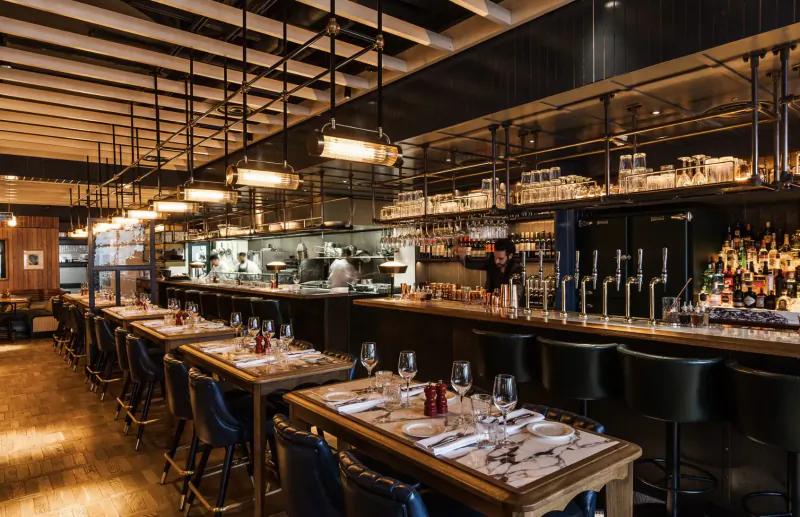 Modern industrial restaurant interior at Battersea Power Station with sleek bar, wooden tables set for dinner, and open kitchen.