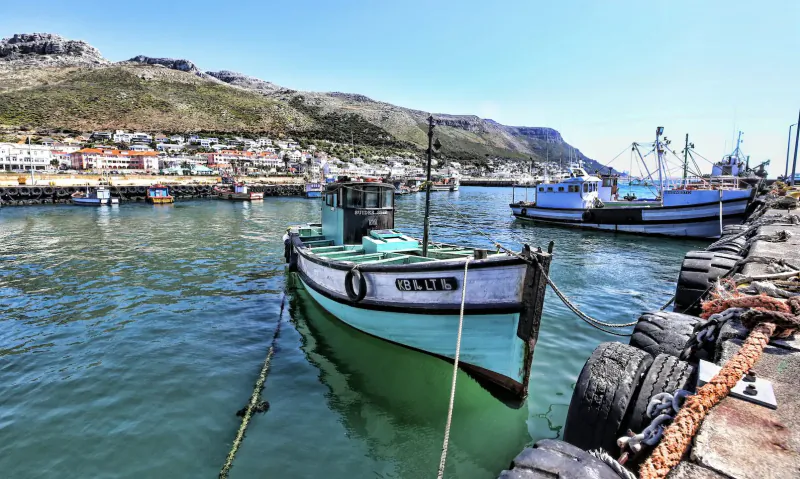 Green and white fishing boat 'Suider Ster' moored in Kalk Bay harbour, Cape Town, with blue boats, pier, and mountain backdrop.