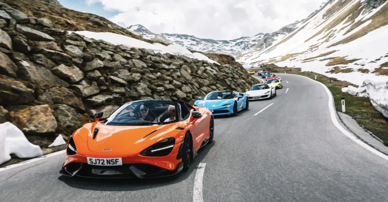 Orange McLaren 720S leads blue McLaren on snowy mountain road, plate SB72 RPO.