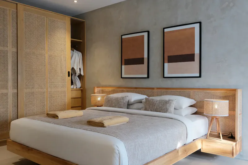 Cozy bedroom in beachfront villa with wooden rattan bed, folded towels, lamps, open wardrobe, and abstract art on textured wall.