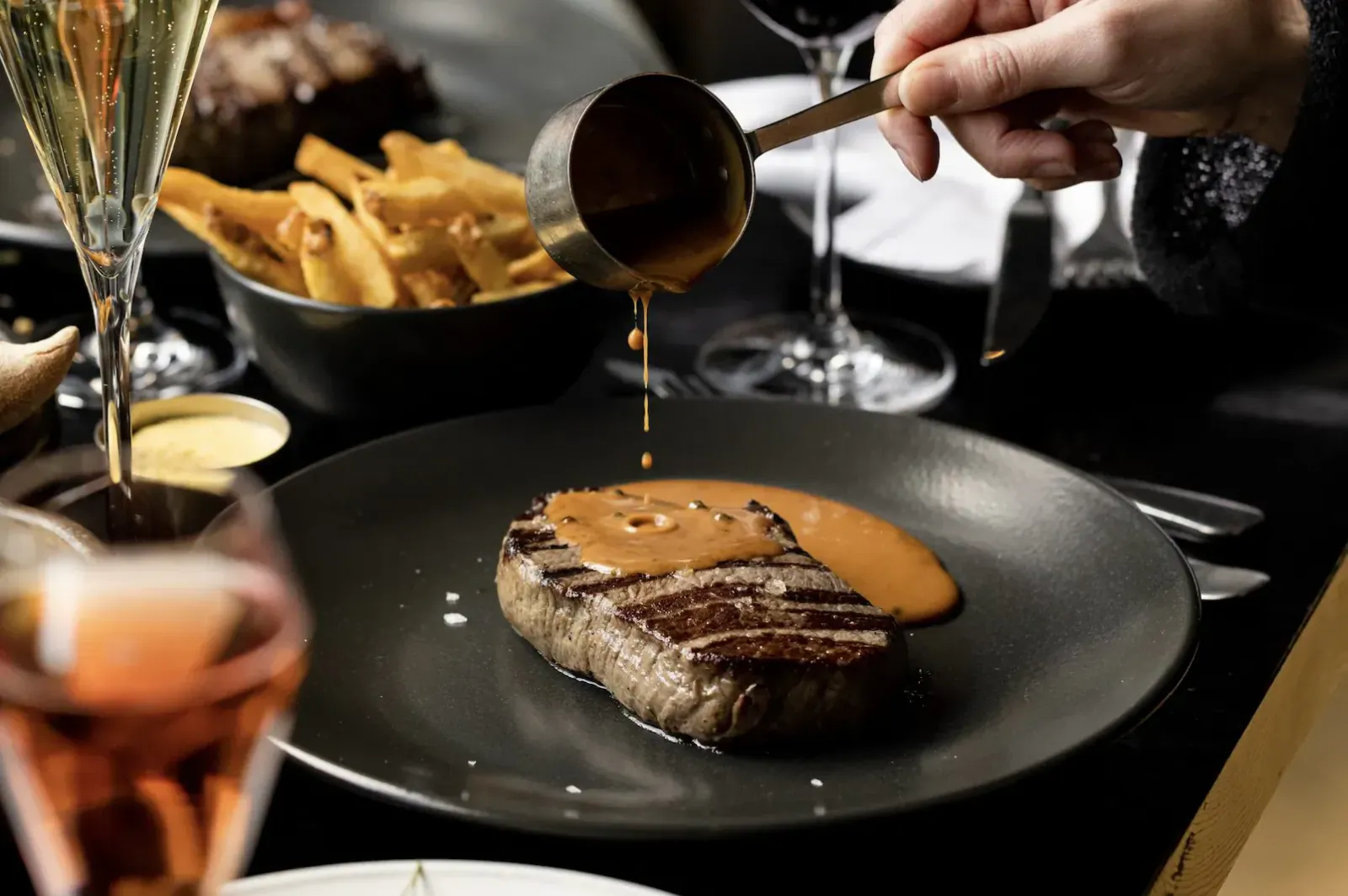 Hand pouring sauce from ladle over grilled steak on plate, with fries, wine glasses at Gaucho restaurant table