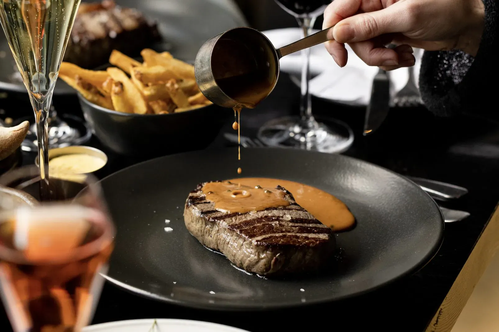 Hand pouring sauce from ladle over grilled steak on plate, with fries, wine glasses at Gaucho restaurant table
