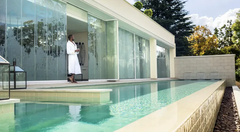 Woman in white robe stands by infinity pool at modern hotel overlooking Johannesburg cityscape.
