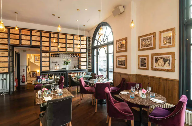 Elegant 28°-50° Chelsea dining room with purple velvet chairs, wooden tables set with wine glasses, wine racks, and arched windows.