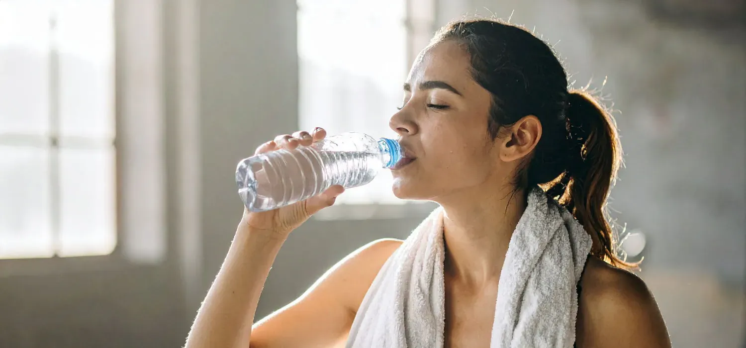 Athletic woman with ponytail and towel drinks from a clear water bottle in sunlit room