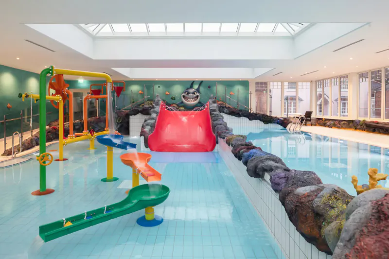 Indoor children's pool at Four Seasons Hampshire with colorful water slides, shark wall, and rock features under skylight.