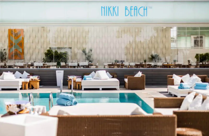 Nikki Beach Ibiza poolside lounge with white daybeds, towels, drinks, and 'Nikki Beach' sign above tiled wall